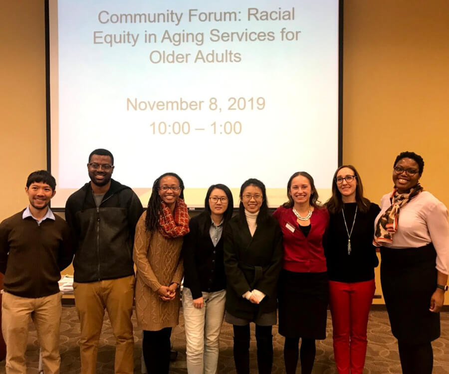 Dr. Tetyana Shippee with her team and community partners standing in a presentation room in a line in from of the podium and screen, which reads, “Community Forum: Racial Equity in Aging Services for Older Adults, November 8, 2019, 10:00 – 1:00