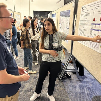 Ayesha standing in front of her poster wearing black pants and a white top pointing to a chart she is explaining to a judge who is looking at the chart