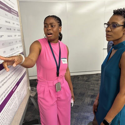 Breanna standing in front of her poster wearing pink pantsuit pointing to a chart she is explaining to Faustine who is looking at the chart