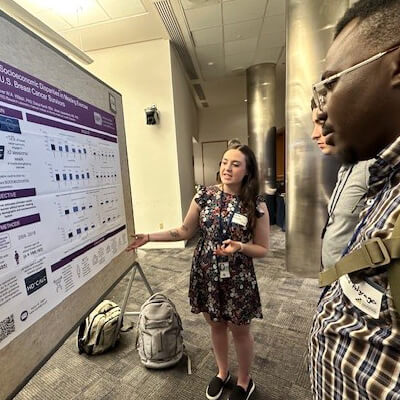 Katie standing in front of her poster wearing a black floral dress explaining her research to two researchers who are looking at her poster