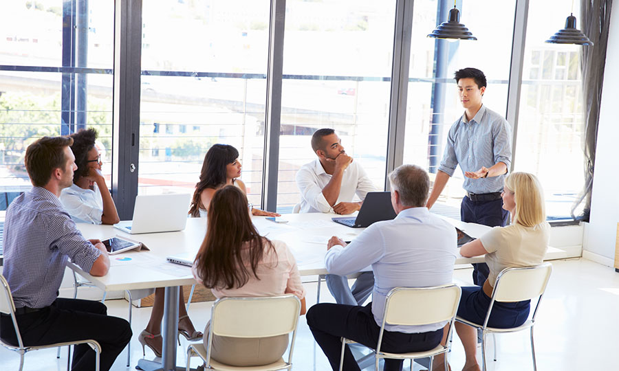 People of different genders, races and ethnicities in a meeting, seated, listening to a man standing at the head of the conference table