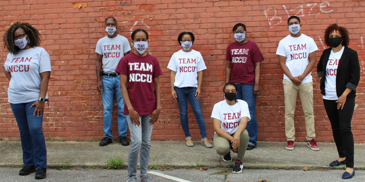 Eight scientists with masks on standing in front of a brick wall