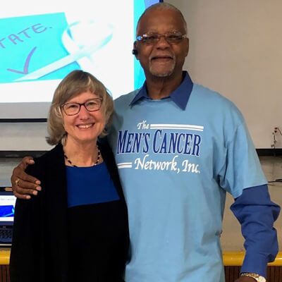 Photo: Dr. Ann Hamilton and Mr. Freddie Muse, Jr. each with an arm on the others’ shoulders, in front of a projector screen at a prostate health event