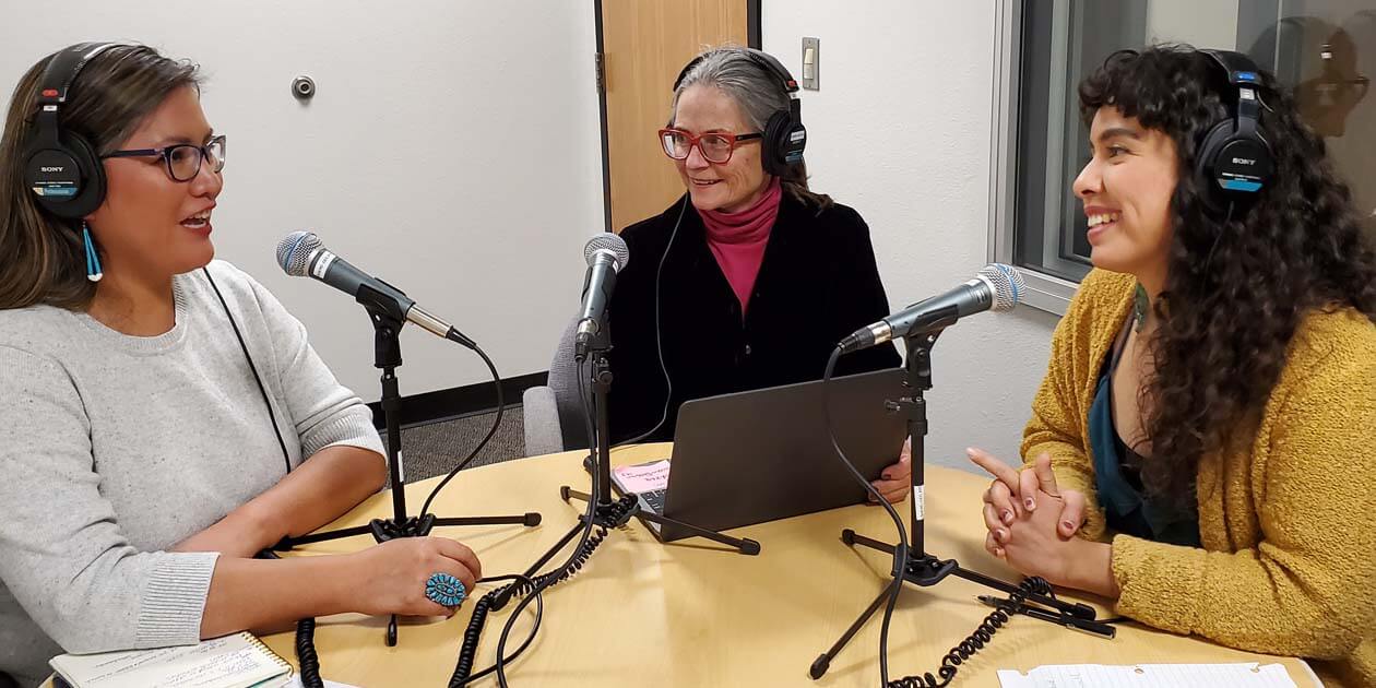 Three women sitting at a table recording a podcast.
