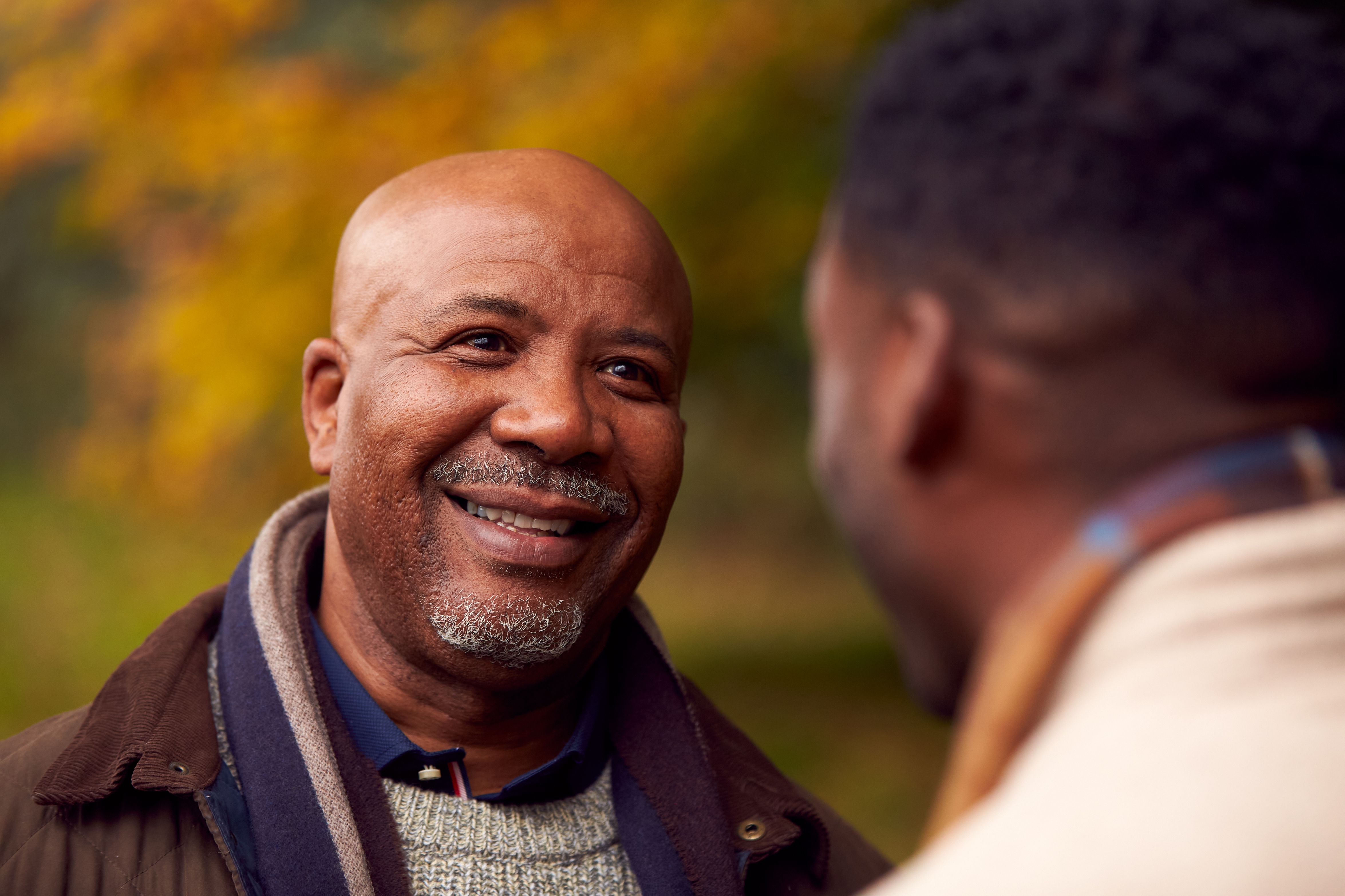 A Black father talking with his adult son, making direct eye contact, a caring smile engaging laugh lines. His son’s partial profile hints at a smile