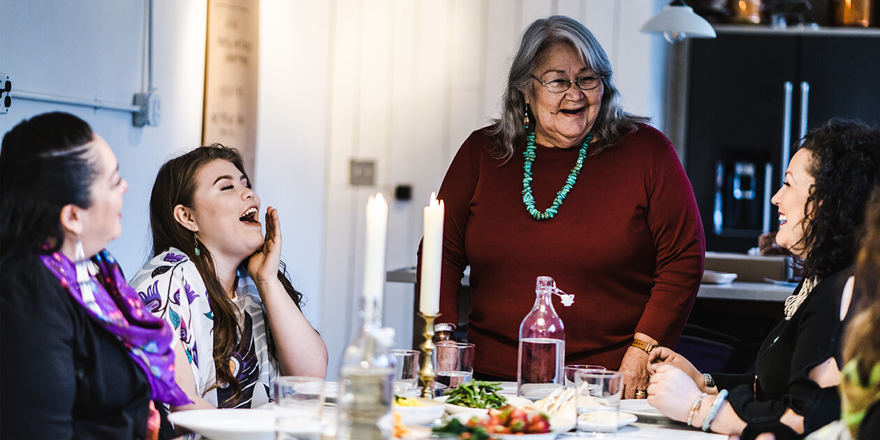 A group of Native American women around a table set with food and 2 lit candles. 3 are seated; an elder wearing a turquoise necklace stands at the head.