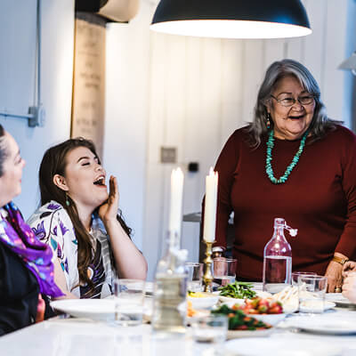 A group of Native American women around a table set with food and 2 lit candles. 2 are seated; an elder wearing a turquoise necklace stands at the head.