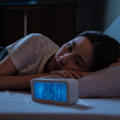 A young woman lying on a bed in a dimly lit room with her eyes open. The clock in front of her reads 2:50.