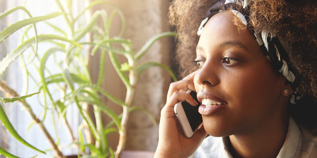 Photo: A young Black woman talking on the mobile phone she’s holding to her ear. Natural light, a green plant and light curtain are behind her