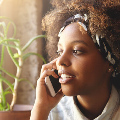 Photo: A young Black woman talking on the mobile phone she's holding to her ear. Natural light, a green plant and light curtain are behind her