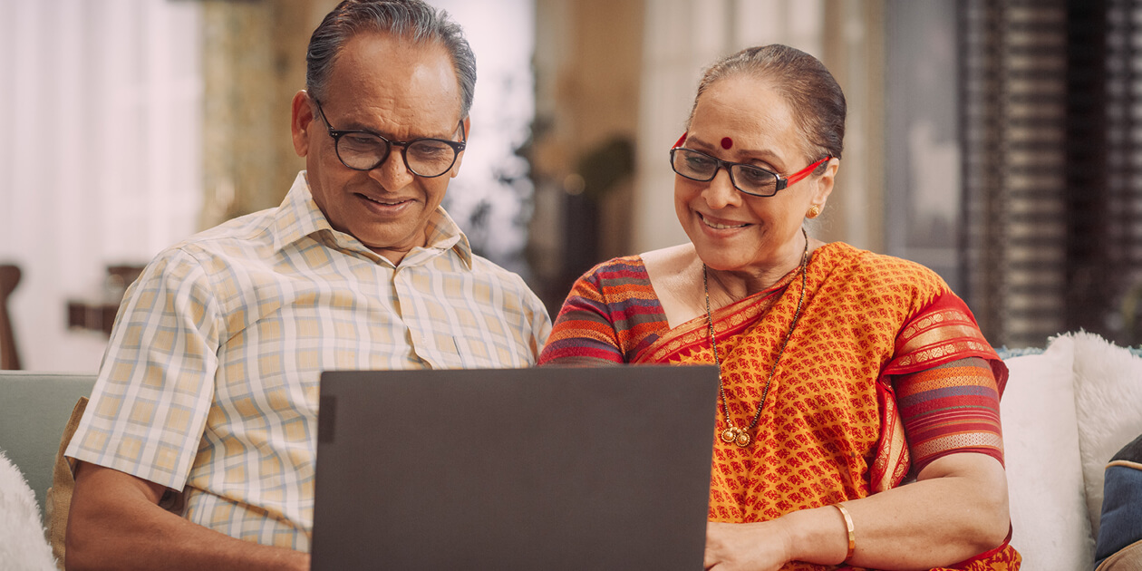 Older Indian couple, he in a collared shirt, she in an orange patterned sari, sit together at home on a couch looking at a laptop screen