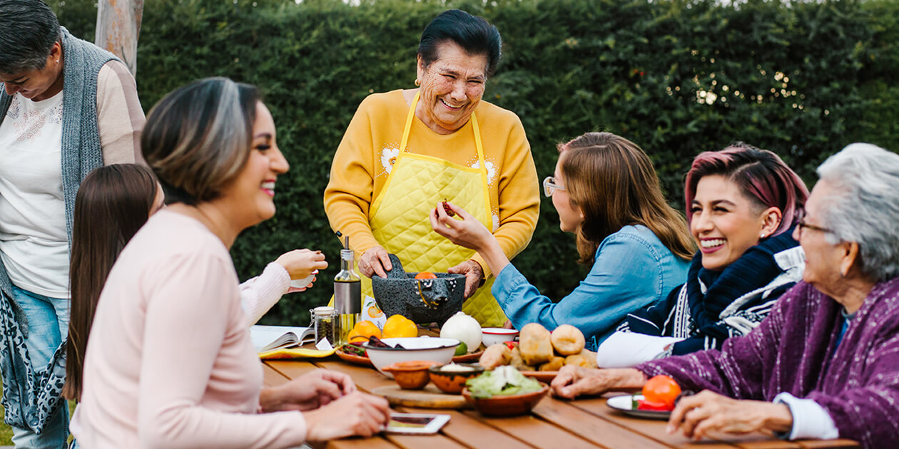 A group of smiling women of Latino descent interact outdoors around a table with dishes of food; 1 stands at the head, working a mortar and pestle