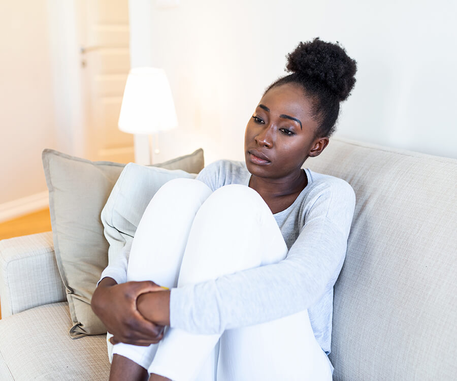 A Black woman sits alone on a couch, clasping her knees to chest, staring vacantly forward