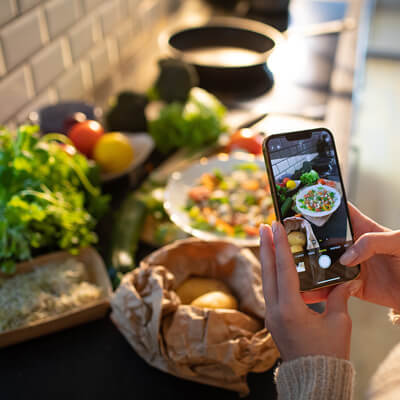 Photo of a woman holding a cell phone taking pictures of food on her kitchen counter in various states of preparation