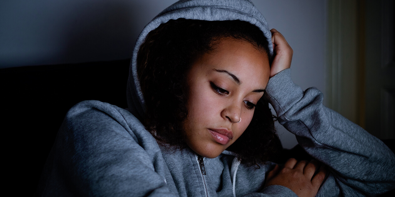 Photo: A Black teenage girl in a blue hoodie sits in a darkened room, her head resting against her right hand, eyes downcast