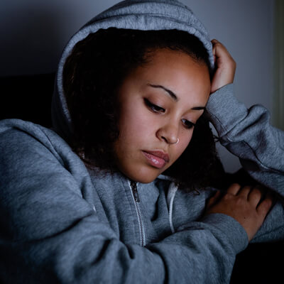 A Black teenage girl in a blue hoodie sits in a darkened room, her head resting against her right hand, eyes downcast